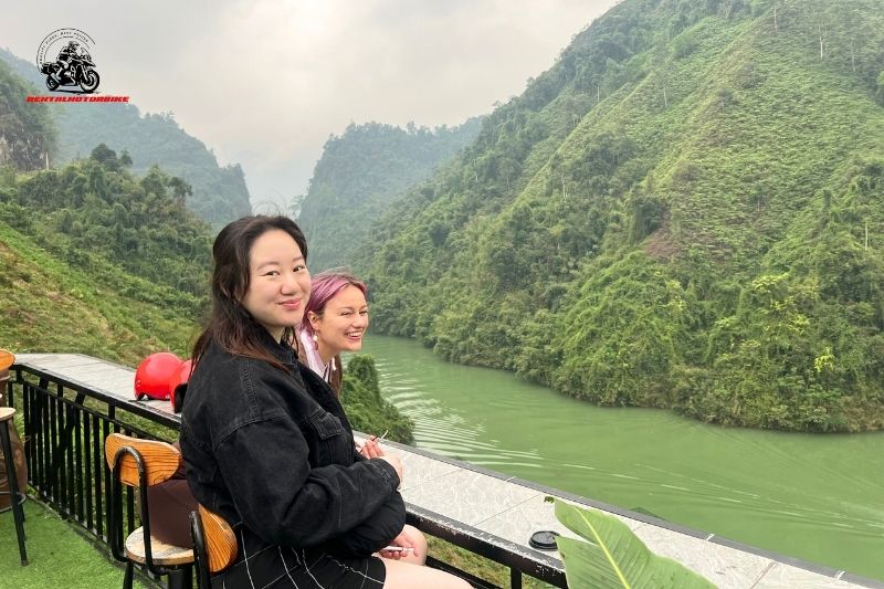 Two women sitting on a terrace overlooking a green river and mountains