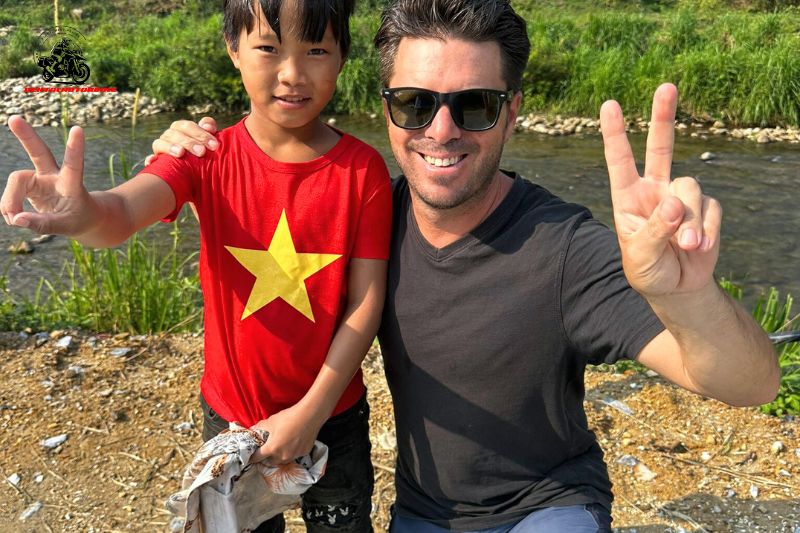 A man and a young boy, both smiling and showing peace signs, standing by a river in a rural setting