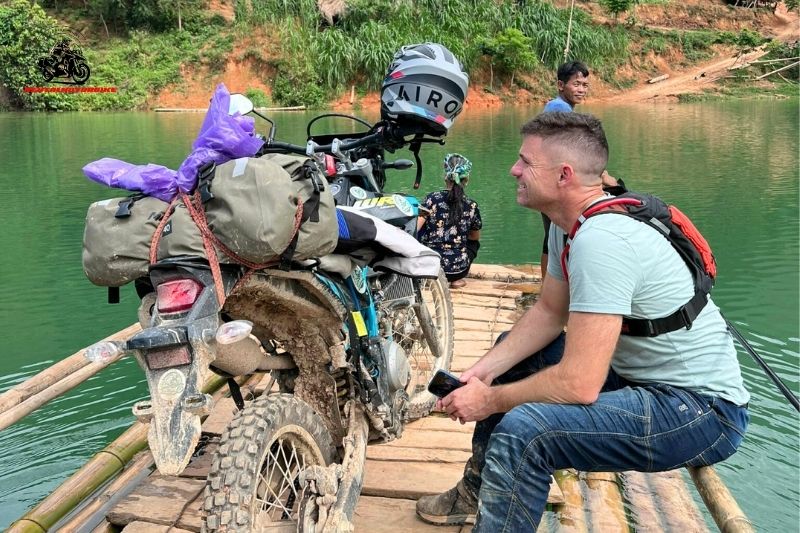 Foreigner sitting beside motorbike on raft crossing a green river