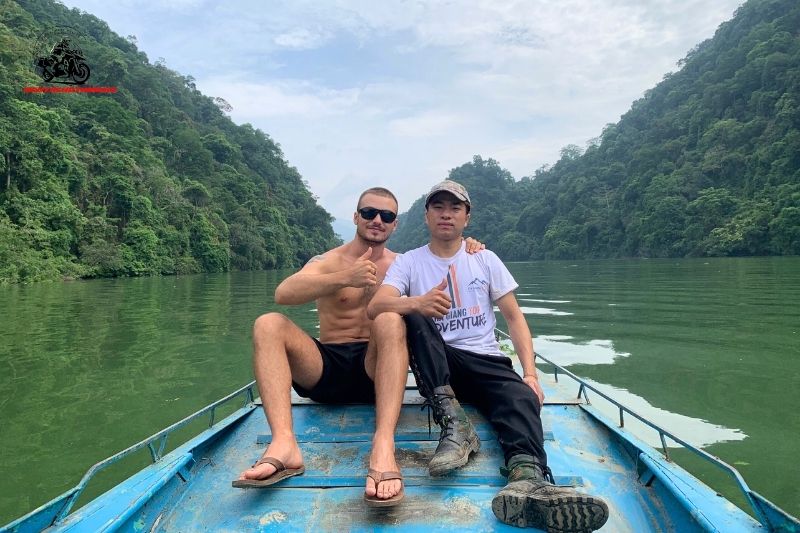 Two men sitting on a blue boat in a forest-lined river or lake