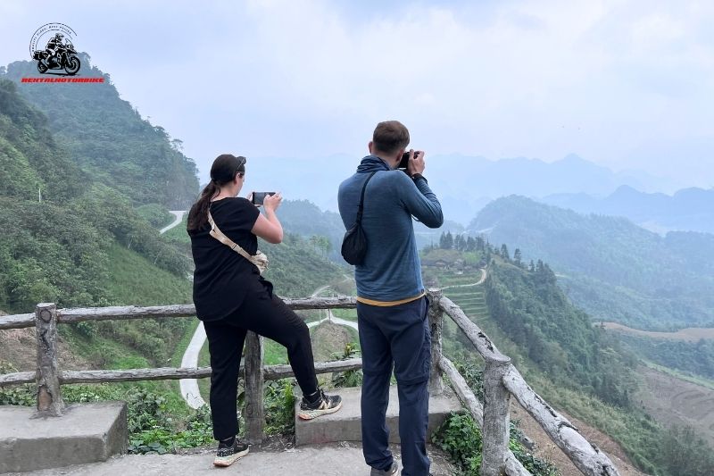 Two tourists taking photos of terraced hills and winding road in Ha Giang